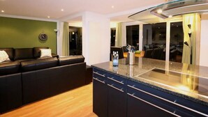 Una sala de estar moderna con un sofá de cuero negro, una zona de cocina con armarios oscuros y encimera de granito, y un comedor con mesa y sillas de vidrio.