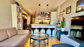 A modern living room with a sofa, armchair, and a glass coffee table. There is a kitchen area with a bar and stools in the background. - Løkka City Apartments (Oslo)