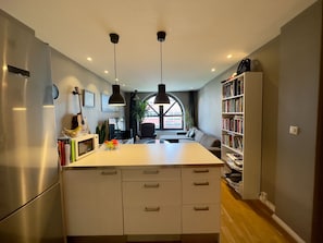 A modern kitchen with a central island, a built-in oven, and a refrigerator. There is a dining area with a sofa and a bookshelf filled with books. - Løkka City Apartments (Oslo)