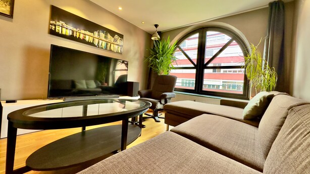 A modern living room with a large flat-screen TV, a glass-top coffee table, a comfortable sofa, and a large window with a view of buildings. - Løkka City Apartments (Oslo)