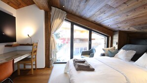 Una camera da letto con soffitto in legno, un letto con biancheria bianca, una scrivania con una sedia e un'ampia finestra che si affaccia su edifici.