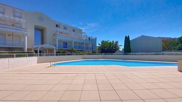 Un área de piscina con suelo de baldosas, un edificio con balcones y un cielo azul despejado.