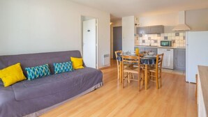 Una sala moderna con un sofá gris, una mesa de comedor de madera y sillas. Se ve una cocina con armarios grises y un microondas al fondo.