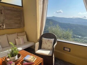 A cozy room with a sofa, a wicker chair, and a table with a plant and books. A window offers a view of distant hills. - Casa Norvegia by Interhome (Valloria)