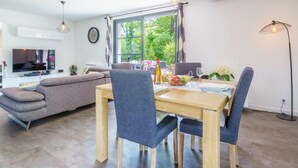 A modern living room with a sofa, dining table, and chairs. There is a TV mounted on the wall, a window with a view of greenery, and a floor lamp. - Chemin du Ruisseau d'Arcangues by Interhome (Anglet)