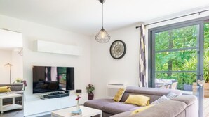 A modern living room with a large flat-screen TV, a sofa with cushions, a small table with a vase, and a large window with a view of greenery. - Chemin du Ruisseau d'Arcangues by Interhome (Anglet)