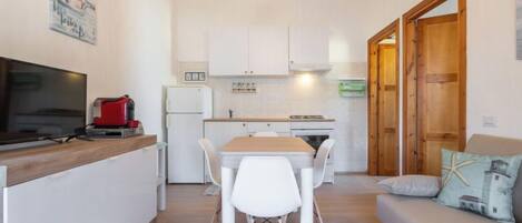 A compact kitchen with white cabinets, a wooden dining table with white chairs, a television, and a coffee machine.