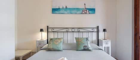 A neatly made bed with two pillows, a bedside table with a lamp, and a painting of sailboats on the wall.