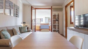 A room with a wooden dining table, a sofa with cushions, a TV on a wooden stand, and a balcony with a view. - Il Faro Blu by Interhome (Villasimius)
