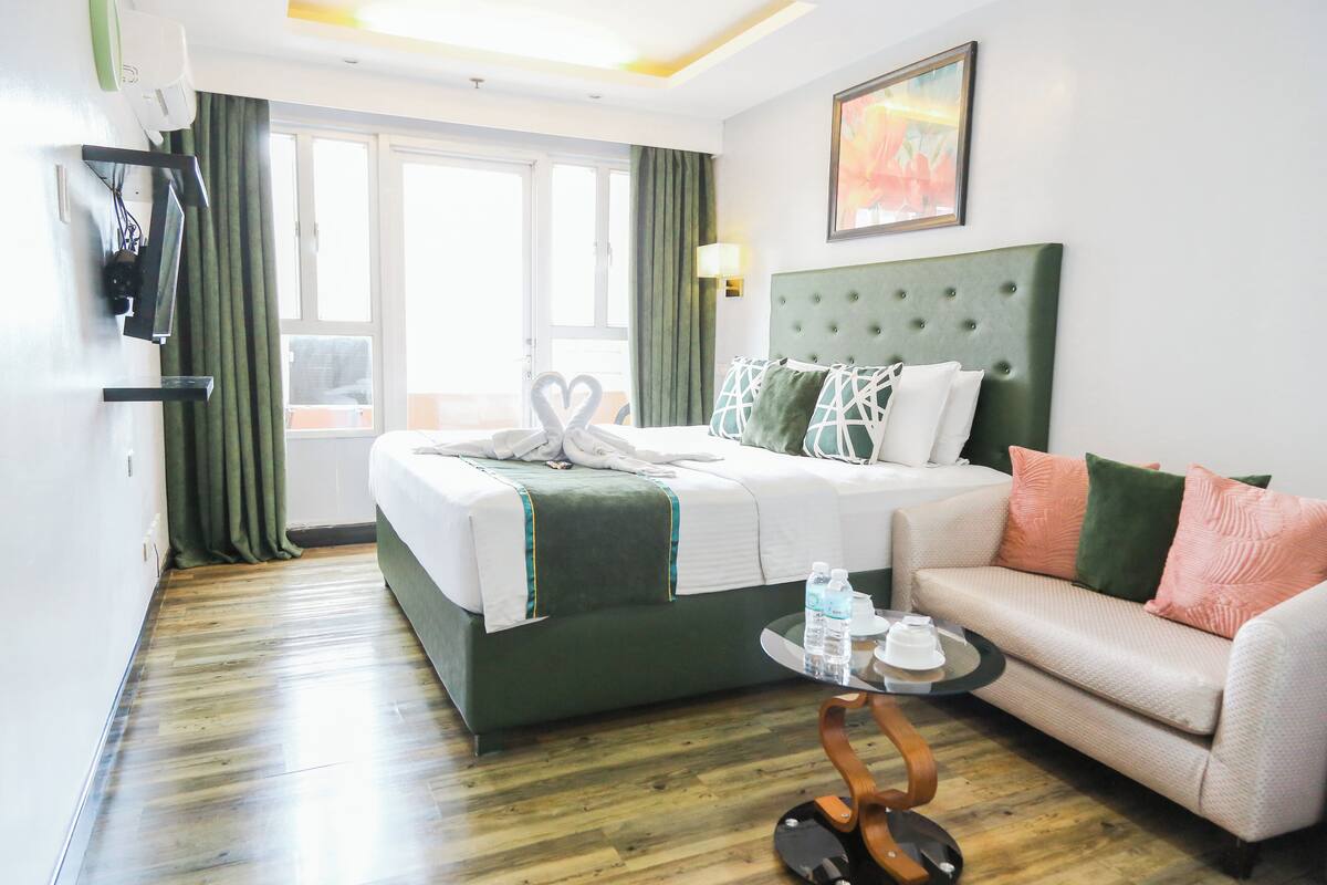 A hotel room with a bed, a sofa, a TV, a window, and a painting on the wall.
