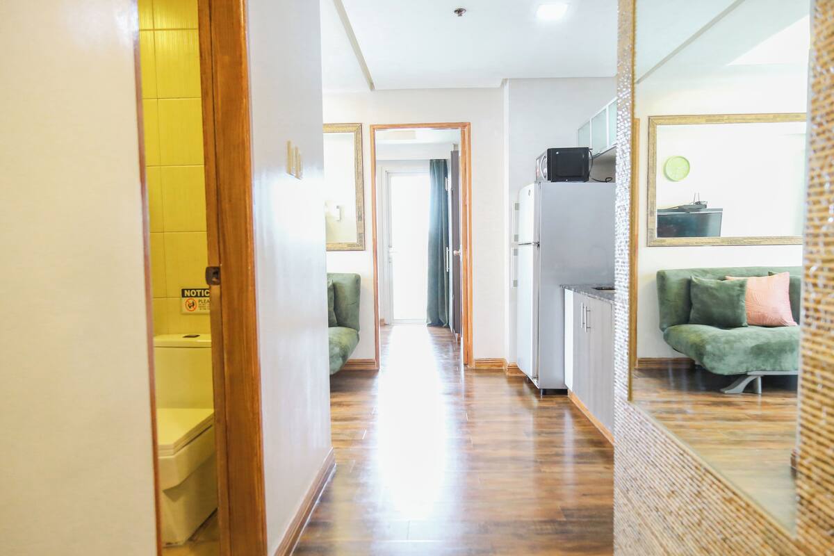 Un pasillo con piso de madera, un sofá verde, un refrigerador blanco y un espejo que refleja la habitación.
