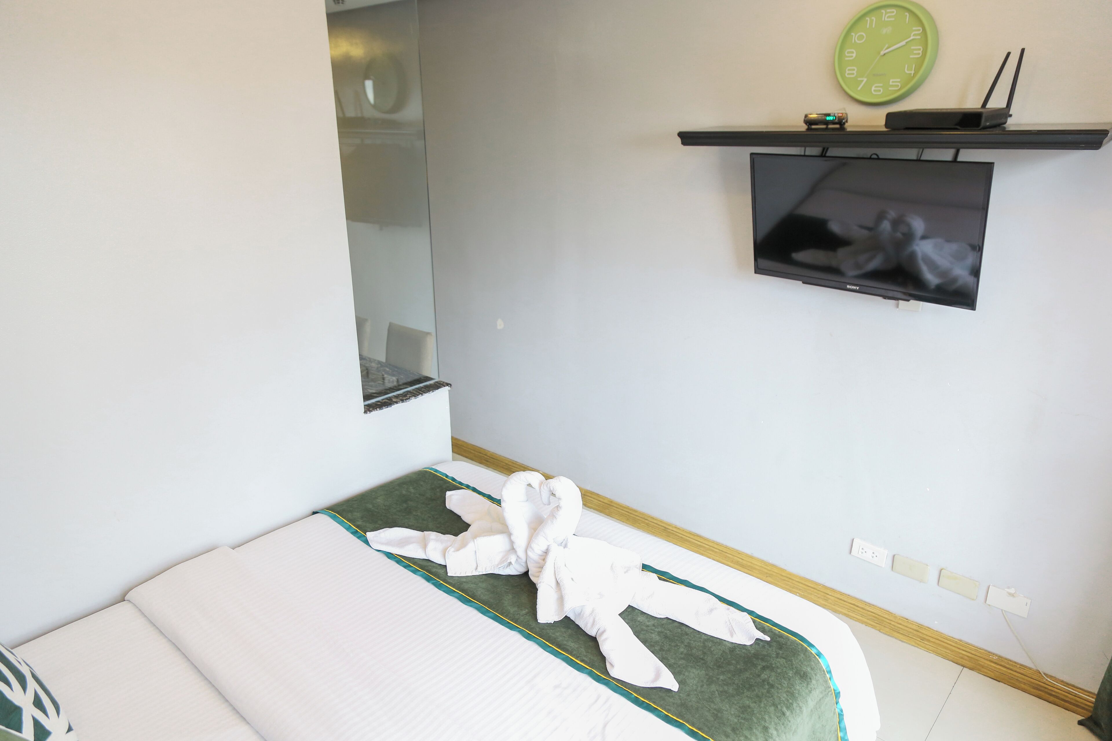 Ein Zimmer mit einem Bett, einem an der Wand montierten Fernseher, einer Uhr und einem kleinen Regal mit einem Gerät.
