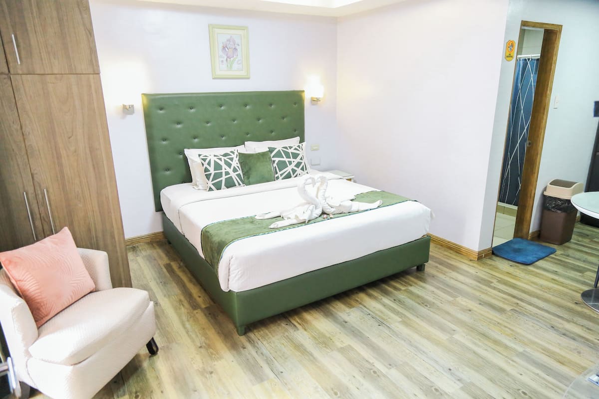Un dormitorio con una cama tapizada de verde, una silla blanca, un armario de madera y piso de madera.
