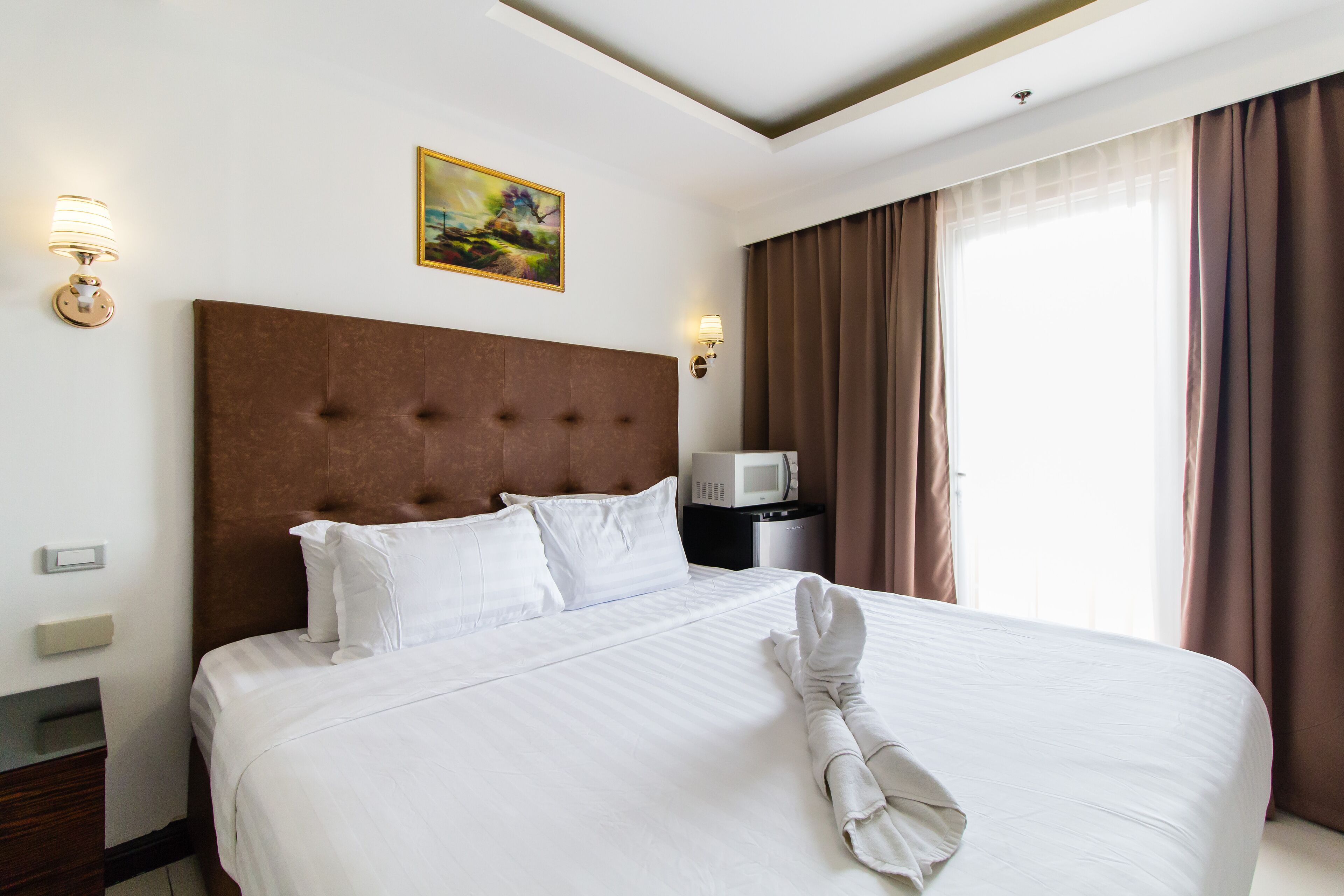 A neatly made bed with white linens and a folded towel, a brown upholstered headboard, a wall-mounted light, a painting, and a window with curtains.