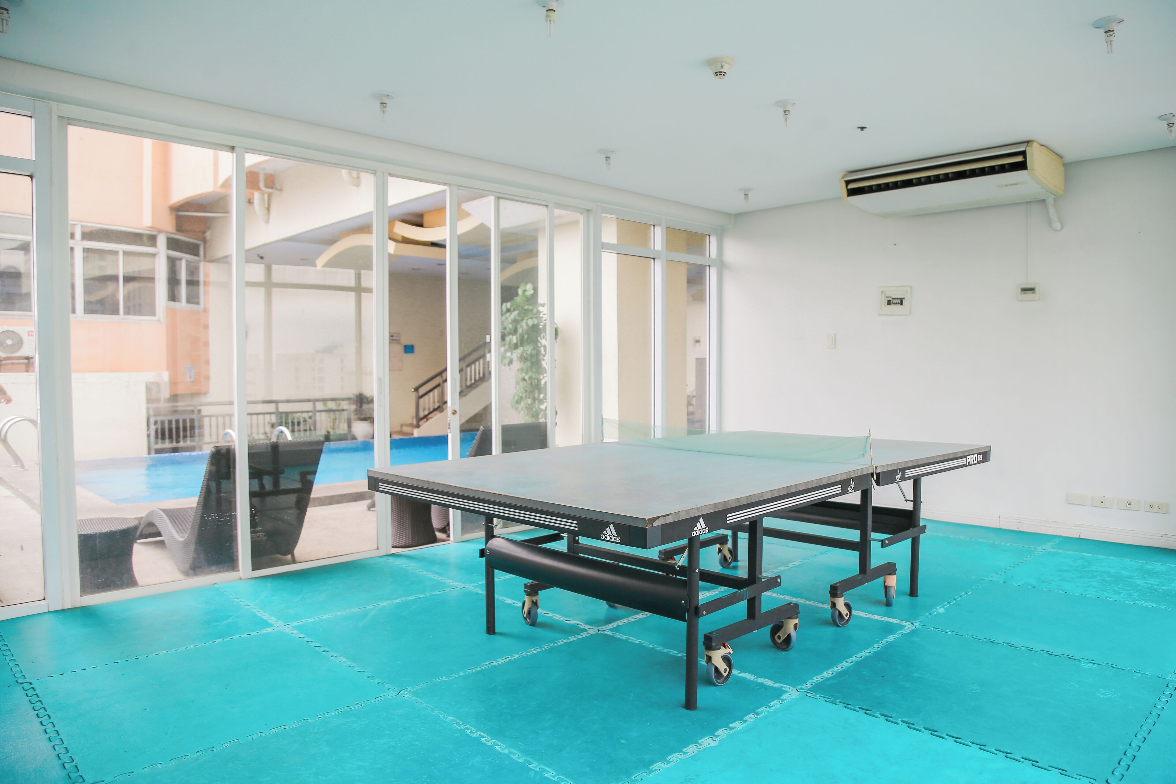 A ping pong table in a room with a blue floor, large windows, and an air conditioning unit on the wall.