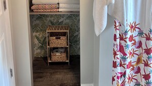 Een badkamer met een douchegordijn met een tropisch patroon, een handdoekhouder met een witte handdoek, en een plank met netjes opgevouwen handdoeken en manden.