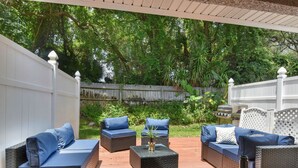 Eine Terrasse im Freien mit Korbmöbeln, darunter Sofas und ein Couchtisch, umgeben von einem Holzboden und einem weißen Zaun.