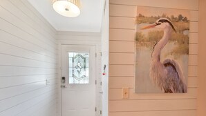 Ein Flur mit weißen Holzwänden, einer Glastür mit geometrischem Muster und einem Gemälde einer Reiher an der Wand.