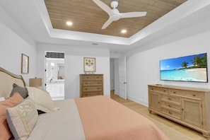 Ein Schlafzimmer mit einem Bett, einem hölzernen Schminktisch, einem Fernseher, einem Deckenventilator und Blick auf einen Strand.