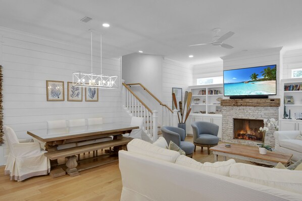 Una sala de estar moderna con chimenea, muebles blancos y un televisor de pantalla plana que muestra una escena de playa.