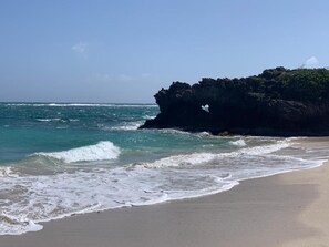 Una playa con agua azul cristalina, olas y un promontorio rocoso.