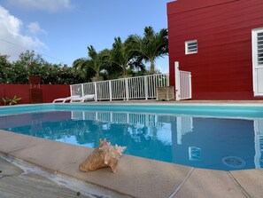 Una piscina con plataforma de hormigón, un edificio rojo y una valla blanca al fondo.