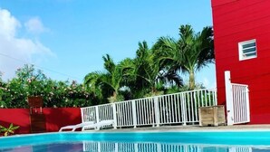 A swimming pool with a white fence, palm trees, and a red building in the background. - Les plages de Macabou (Le Vauclin)