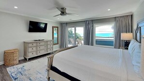 A bedroom with a large bed, a ceiling fan, a TV, a dresser, and a view of the ocean. - Laguna Matata | Patio, Balcony, Private Pool & Hot Tub (Panama City Beach)