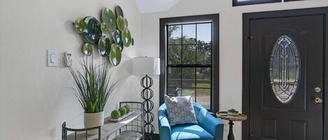 A modern entryway with a blue armchair, a potted plant, a decorative mirror, and a black door with a glass panel.