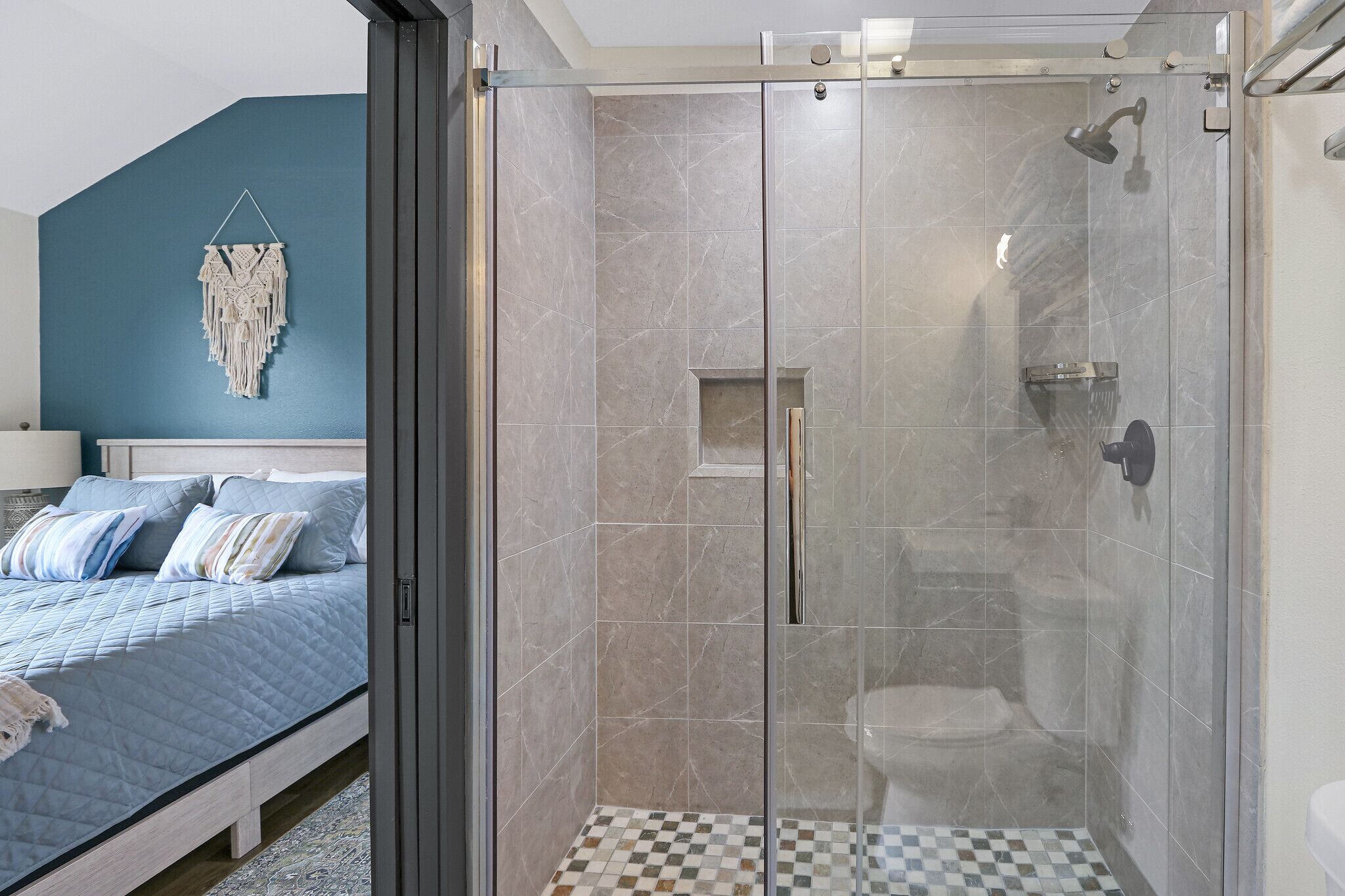 Een slaapkamer met een bed, een nachtkastje en een macramé kunstwerk aan de muur. Grenzend aan de kamer bevindt zich een douche met glazen wanden en een betegelde vloer.