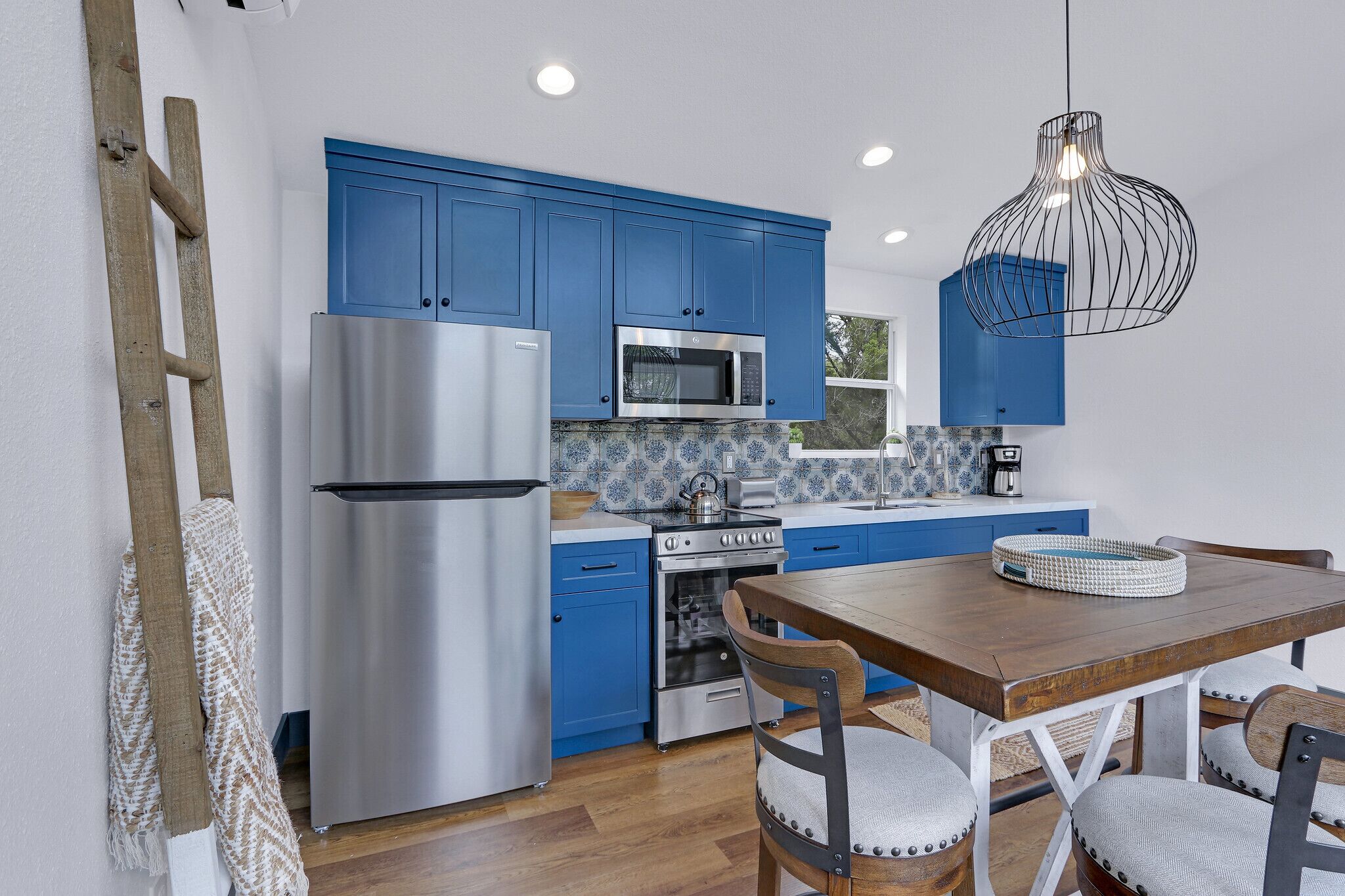 A modern kitchen with blue cabinets, stainless steel appliances, and a wooden dining table.