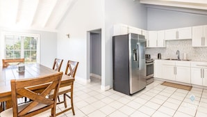 A modern kitchen with white cabinets, a stainless steel refrigerator, and a dining area with wooden chairs and a table. - Lagoon Views, Private Balcony & Near the Beach (Bottle Creek)