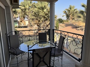 A balcony with a glass table and four chairs, overlooking palm trees and a tiled roof.