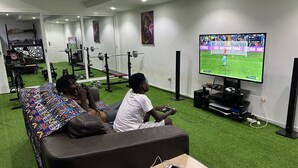 A living room with a large TV displaying a soccer game, a sofa, and a coffee table with a smartphone on it. - Chez Lilie Maison d'hôtes à Saly (Mbour)