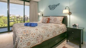 Een slaapkamer met een groot bed, twee bedlampjes, een nachtkastje en uitzicht op groen door grote ramen.