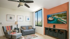 Een woonkamer met een grijze bank, een stoel met een kleurrijke deken, een salontafel, een televisie op een dressoir en een groot raam met uitzicht op de kust.
