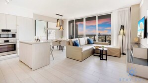 A modern apartment with a kitchen, dining area, and living room. It features a large window with a city view, a sofa, a coffee table, and a TV. - Luxury 1 Br| Front Intracoastal | Balcony |Pools (Hollywood)