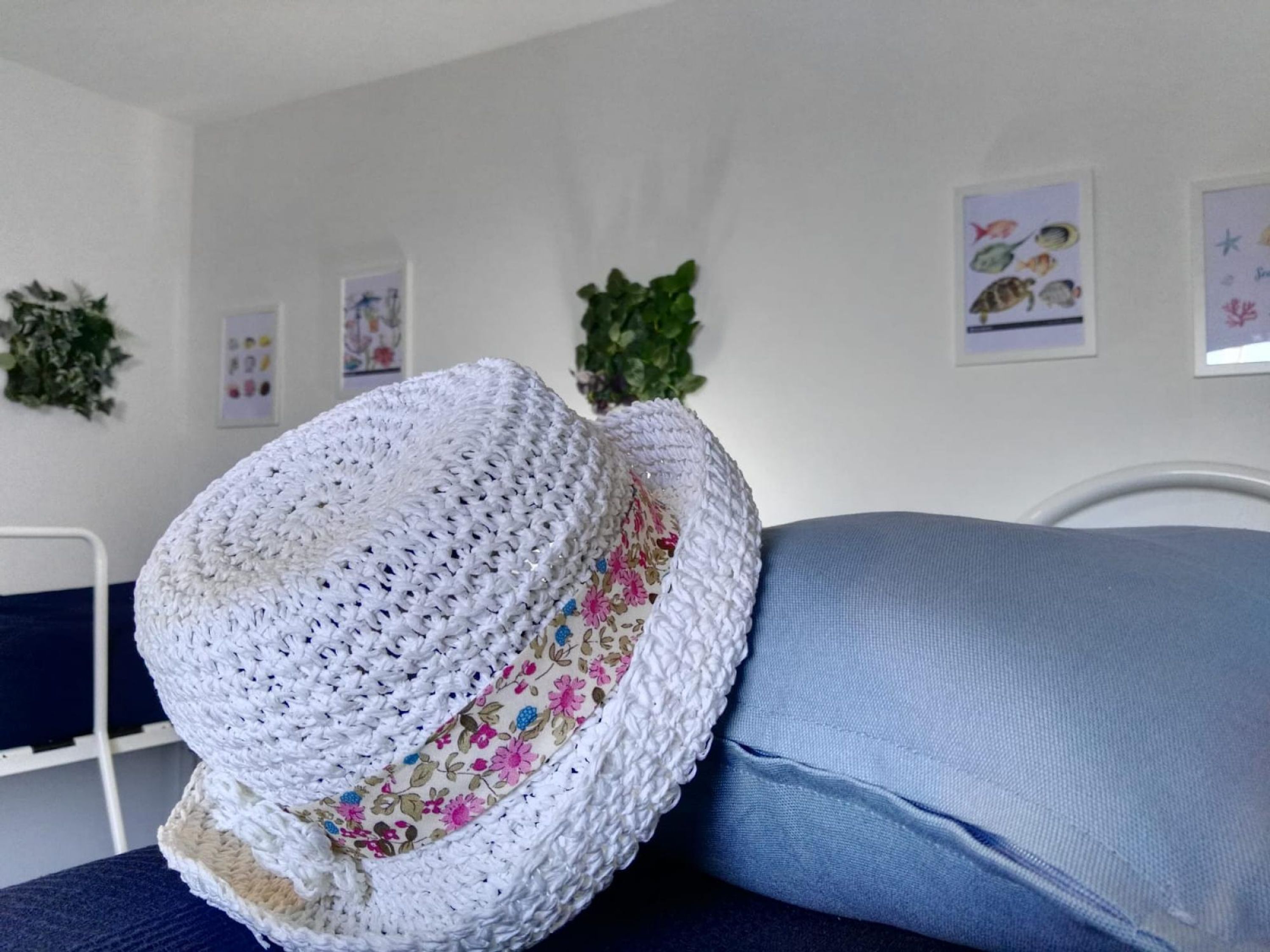Un sombrero blanco de ganchillo con motivos florales sobre un cojín azul en una habitación con paredes blancas y cuadros enmarcados.