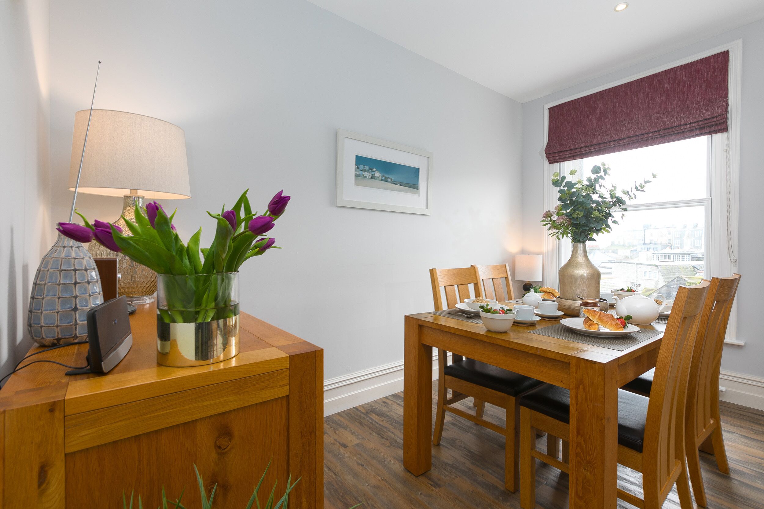 Un coin repas avec une table en bois, des chaises, un vase rempli de fleurs violettes, une lampe et une fenêtre donnant sur des immeubles.