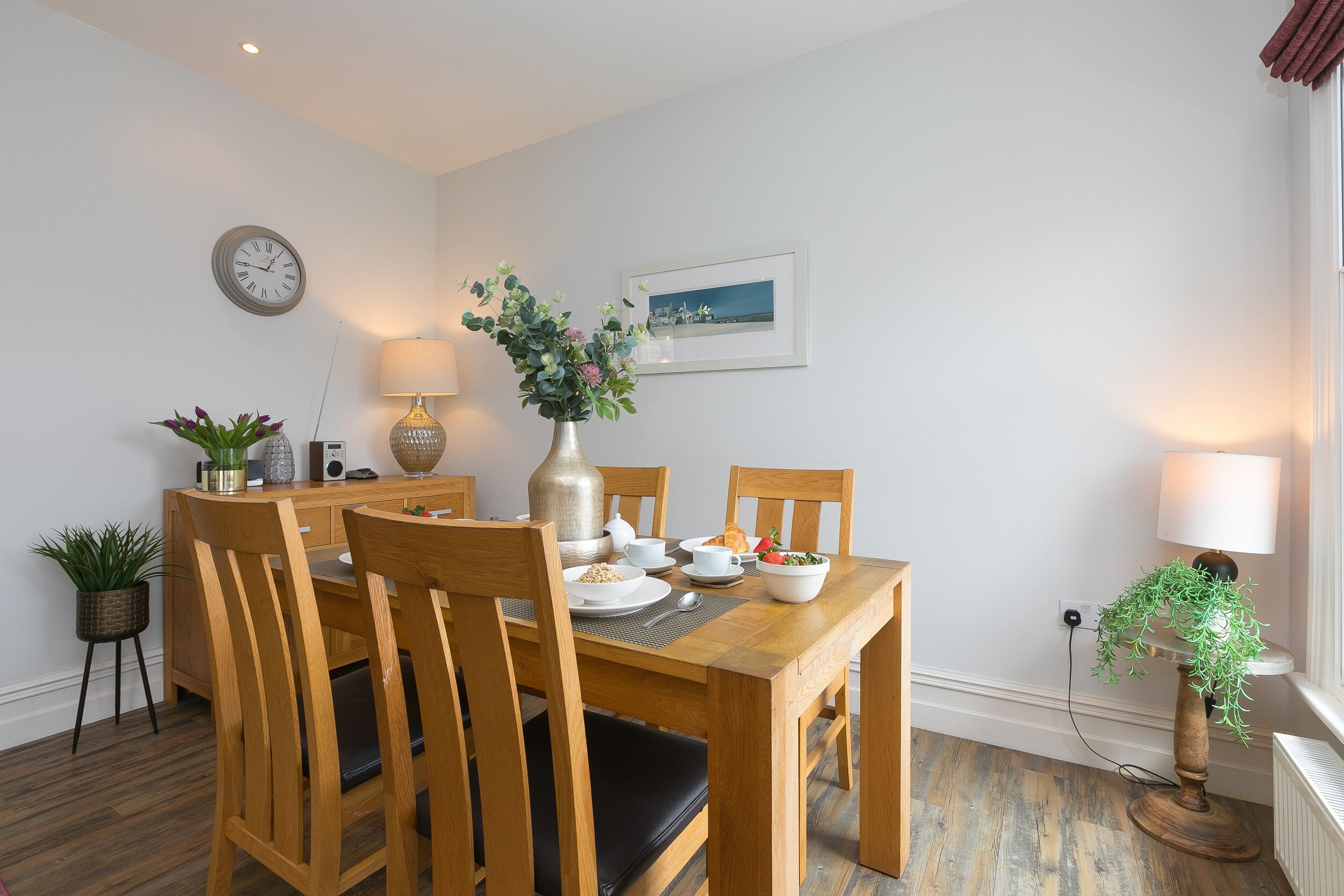A dining area with a wooden table, chairs, a vase with flowers, a clock, and a framed picture on the wall.