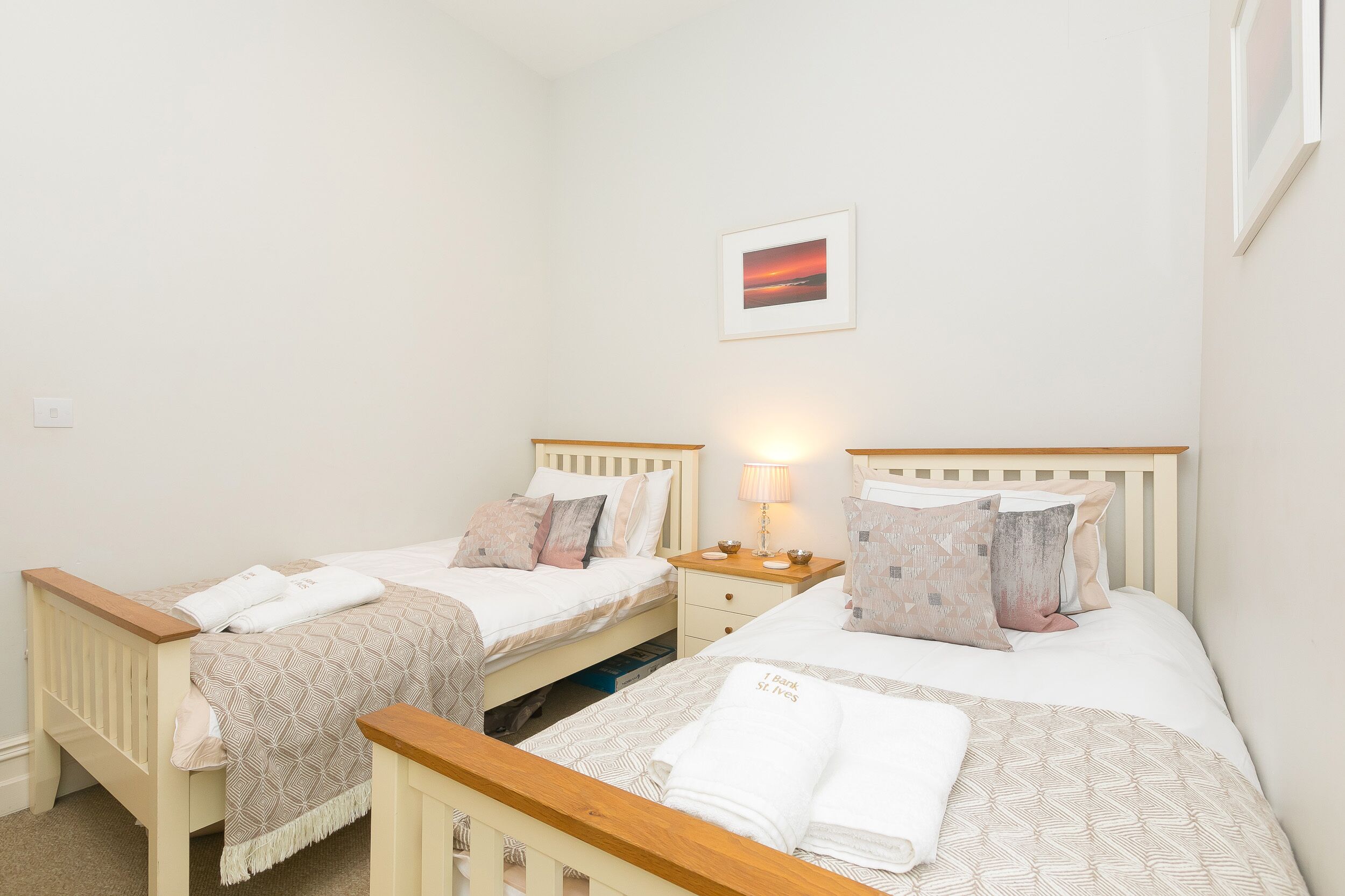 A bedroom with two single beds, each with a wooden headboard, a bedside table with a lamp, and a framed picture on the wall.