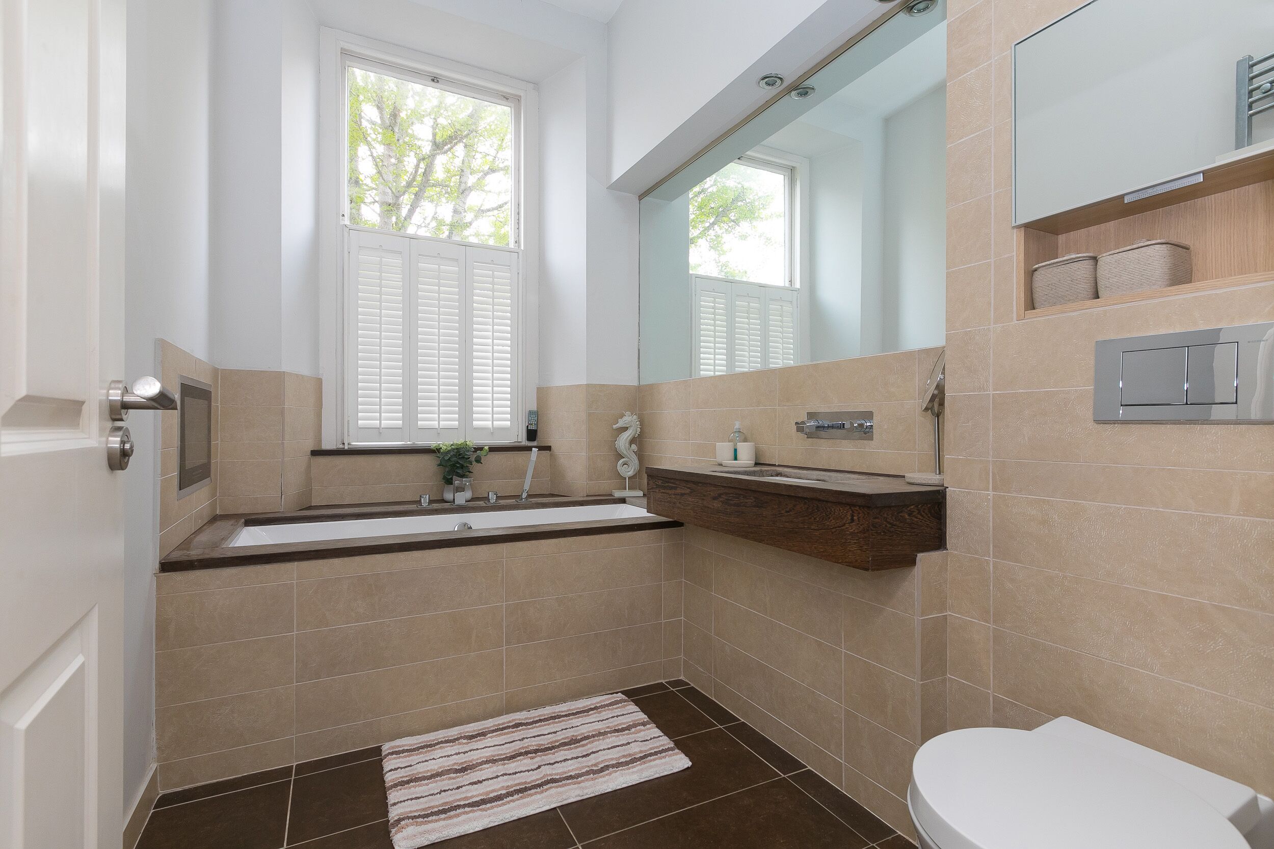 A modern bathroom with a bathtub, sink, and toilet. It features a window, a mirror, and a towel rack.