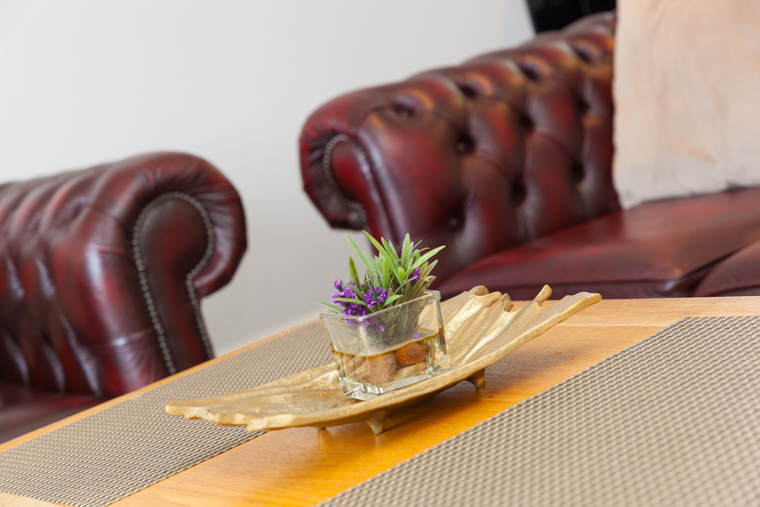 Een eettafel met een glazen vaas met paarse bloemen, een gouden decoratie en een geweven placemat.