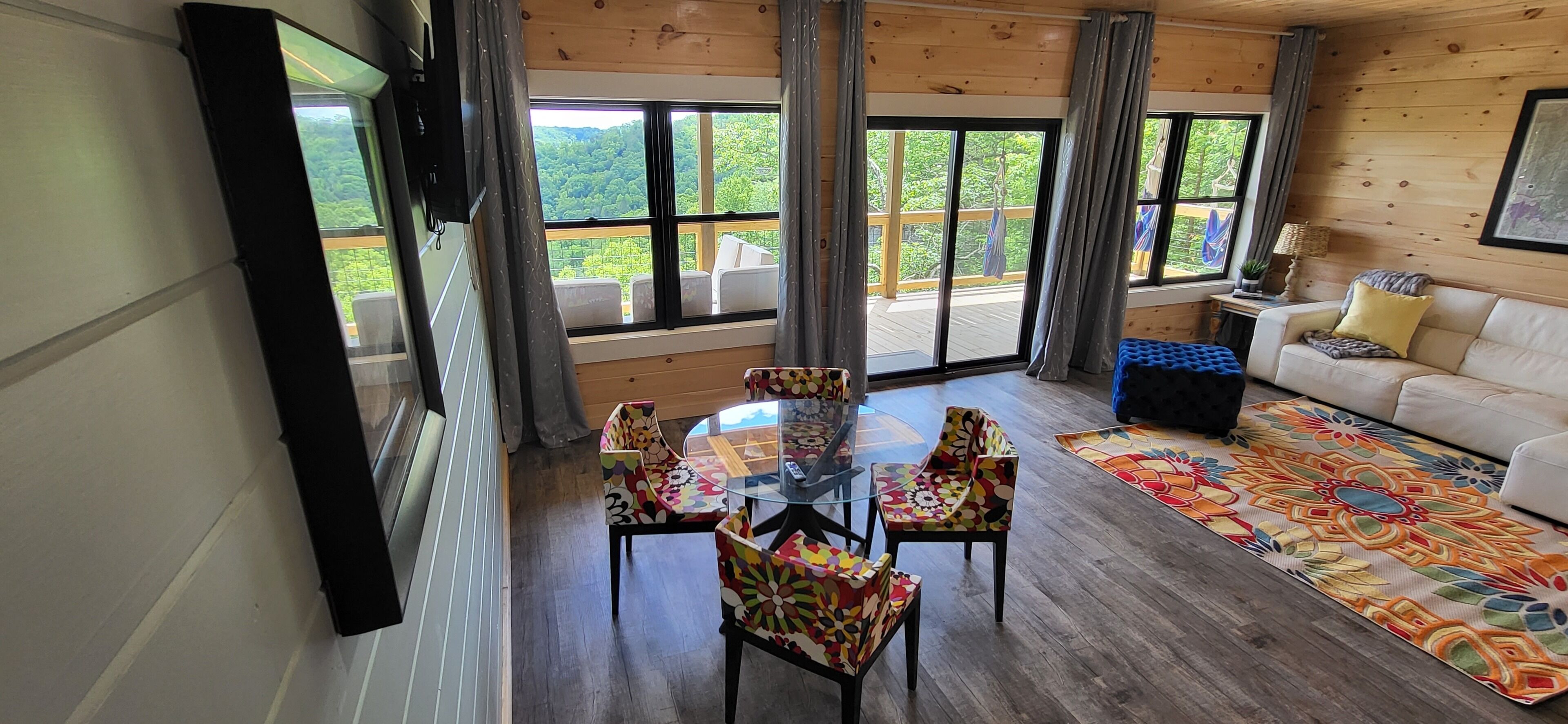 Une pièce spacieuse dotée d’un plancher en bois, d’une table en verre entourée de chaises colorées, d’un canapé blanc et de grandes fenêtres donnant sur une terrasse et un espace verdoyant.