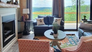 Een gezellige woonkamer met een open haard, een blauwe bank, gestreept fauteuils en een groot raam met uitzicht op de natuur.