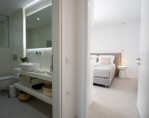 A modern bathroom with a large mirror, a white sink, and a toilet. There is a bed with white bedding in the adjacent room. - Ultimate Luxury Paradise Villa In Paros (Paros)