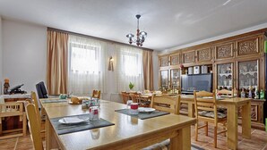 Zona pranzo con tavolo di legno apparecchiato per quattro persone, circondato da sedie. Sopra il tavolo, un lampadario, e a destra un mobile con vetrine.