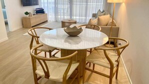 Una sala de estar moderna con una mesa de comedor redonda, cuatro sillas de madera, un sofá, un televisor en la pared y una lámpara de pie.