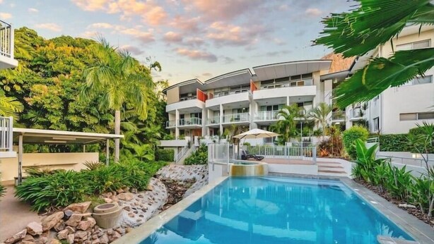 Un hotel moderno con piscina, abundante vegetación y un edificio de varios pisos con balcones.