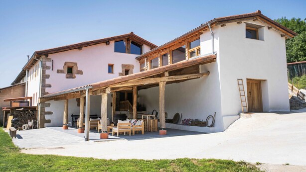 A two-story building with a covered patio area, a wooden staircase, and a small wooden door. - Paorra Baserria (Aia)
